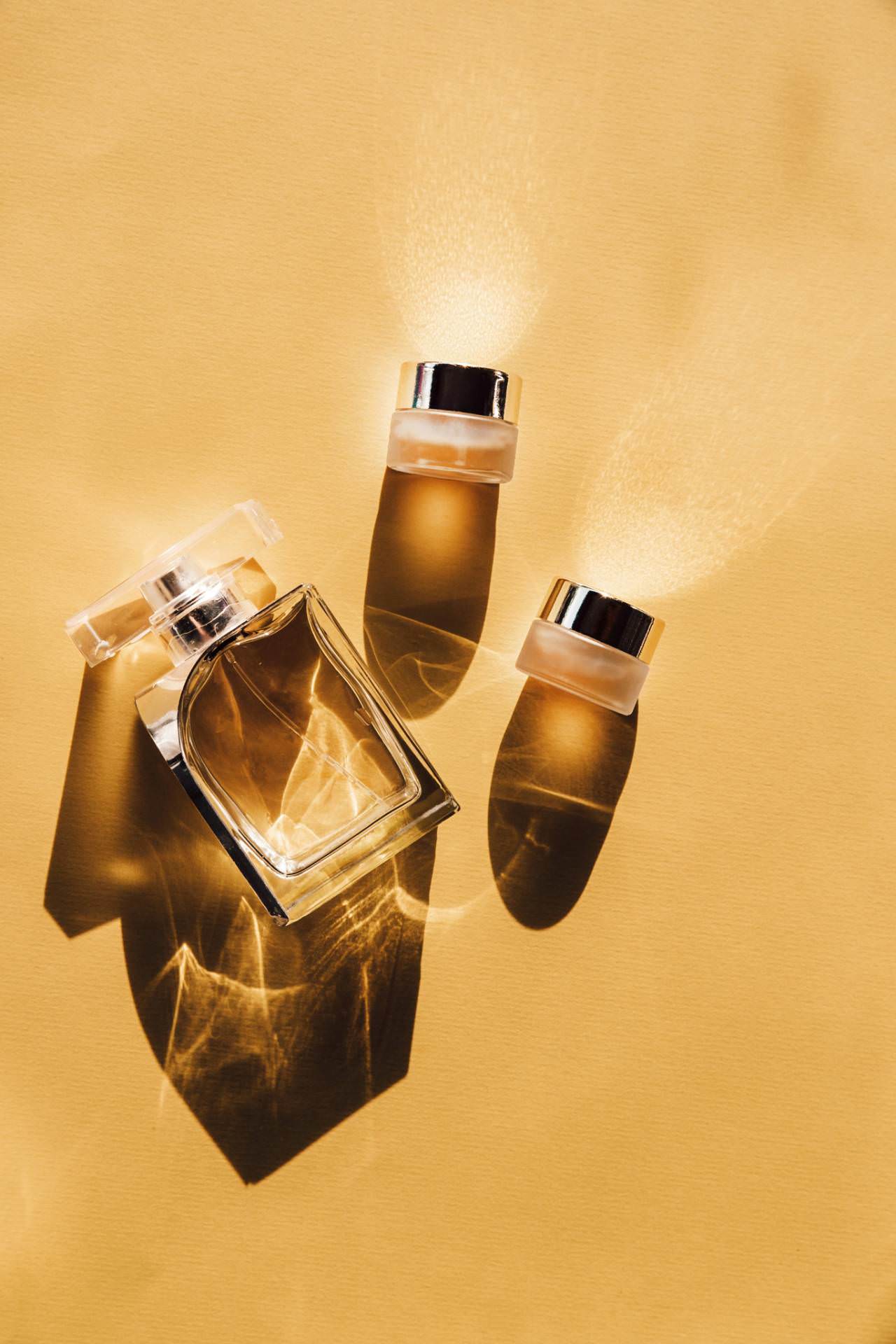 Un flacon de parfum en verre transparent et deux couvercles argentés retirés sont disposés sur une surface jaune clair, projetant des ombres et des reflets lumineux distincts.