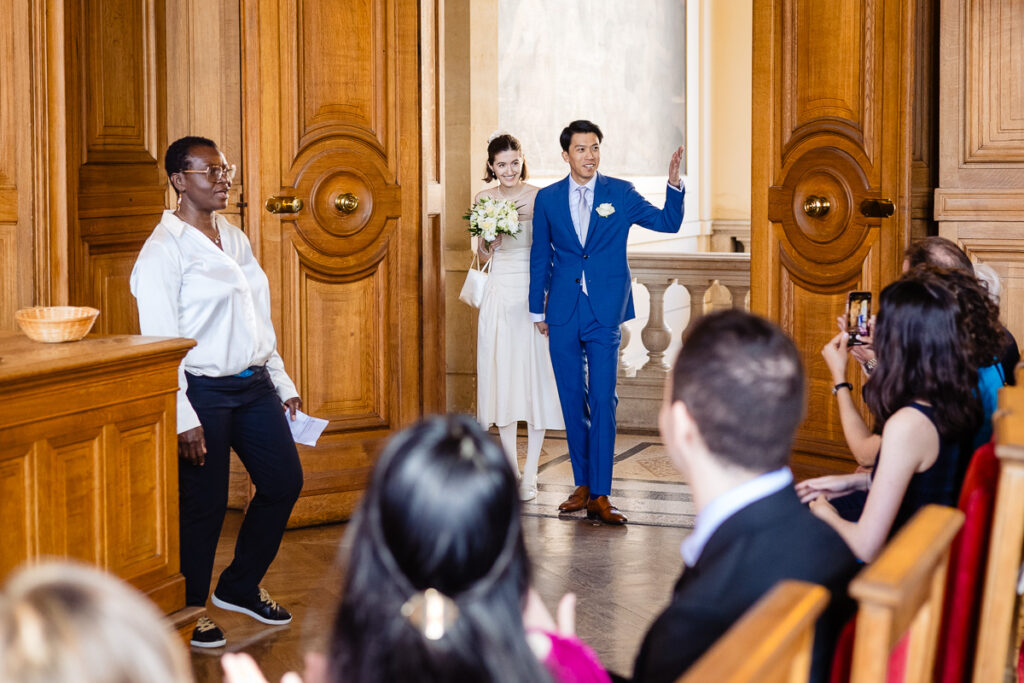 Un couple entre dans une salle pour une cérémonie de mariage, le marié faisant signe de la main, tandis que les invités regardent et qu'un célébrant de mariage se tient à proximité.