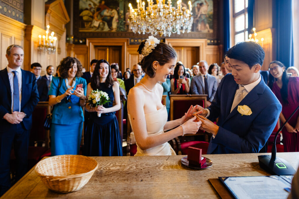 Un couple échange des alliances dans une salle de cérémonie formelle ornée de lustres. Les invités se tiennent autour, certains souriant et prenant des photos. Un panier et un document sont posés sur la table en bois au premier plan.