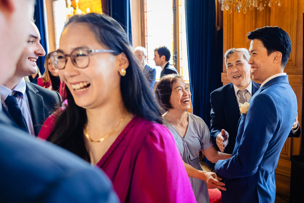 Un groupe de personnes vêtues de tenues formelles rient et discutent ensemble à l'intérieur, avec des hommes en costume et des femmes en robe.