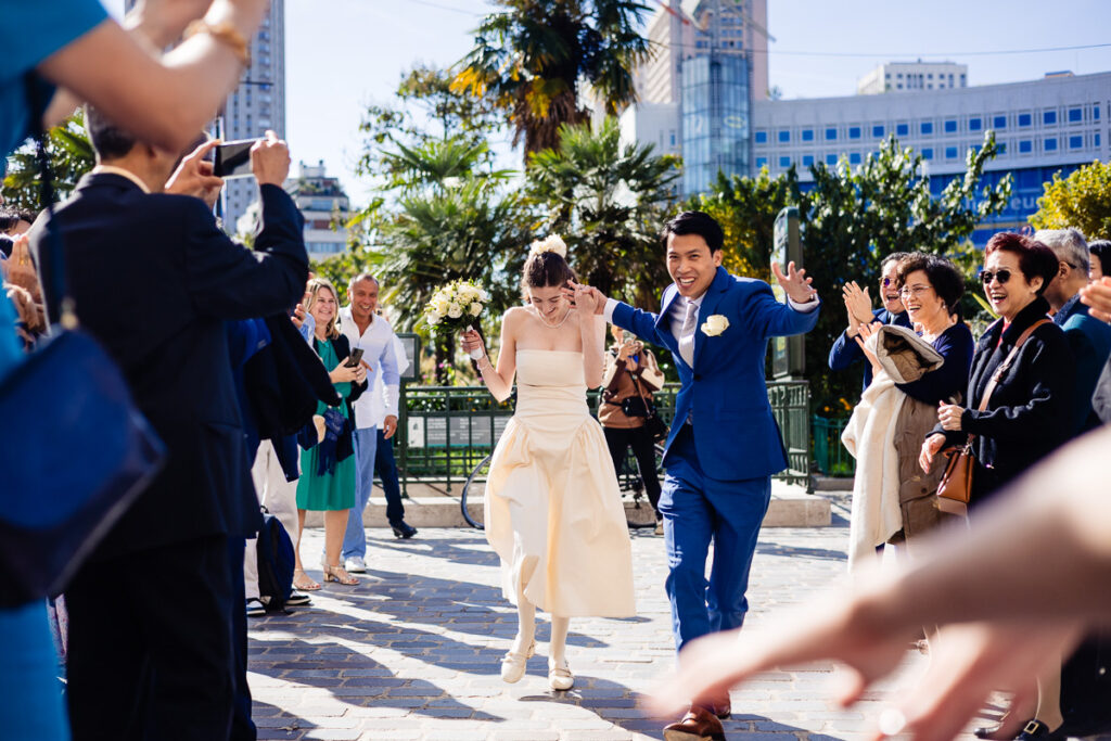 Un couple de mariés marche joyeusement au milieu d'une foule d'invités enthousiastes. Le marié porte un costume bleu et la mariée tient un bouquet.