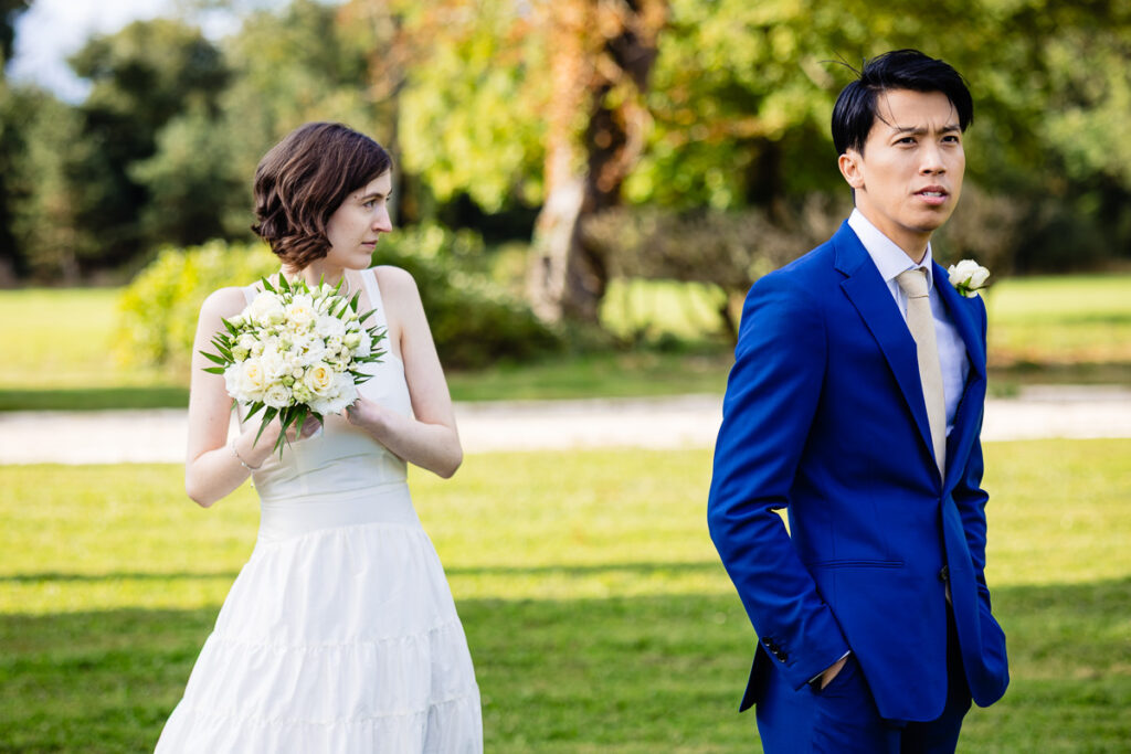 Une mariée en robe blanche tient un bouquet, regardant sur le côté, tandis qu'un marié en costume bleu se tient debout, les mains dans les poches, regardant vers l'avant, dans un décor de jardin.