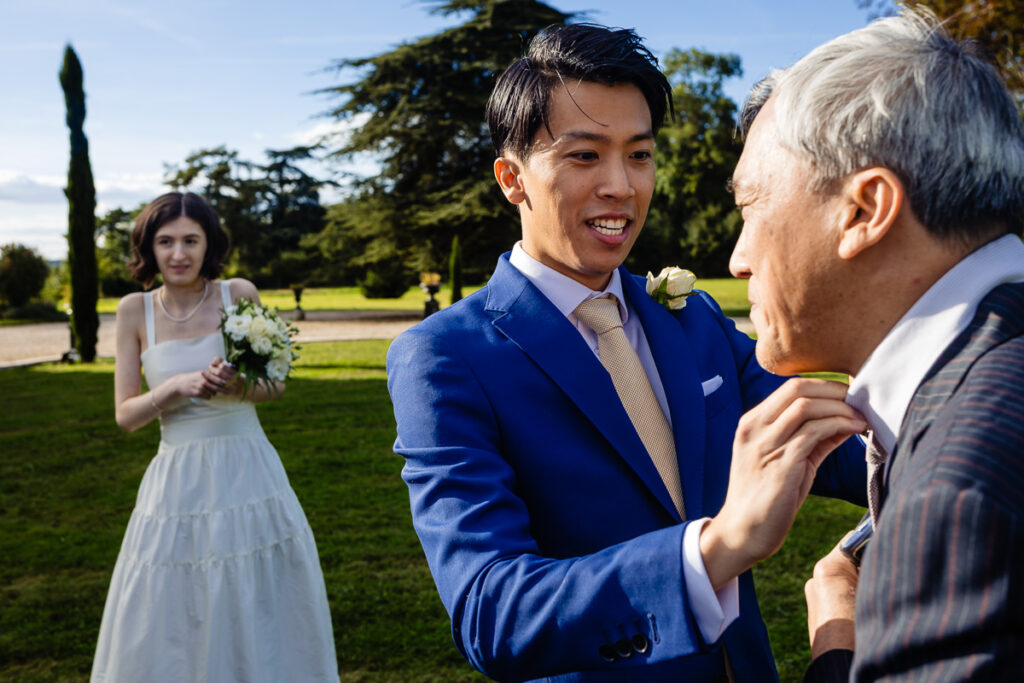 Un marié en costume bleu ajuste le col d'un homme plus âgé à l'extérieur. Une femme en robe blanche tient un bouquet à l'arrière-plan.