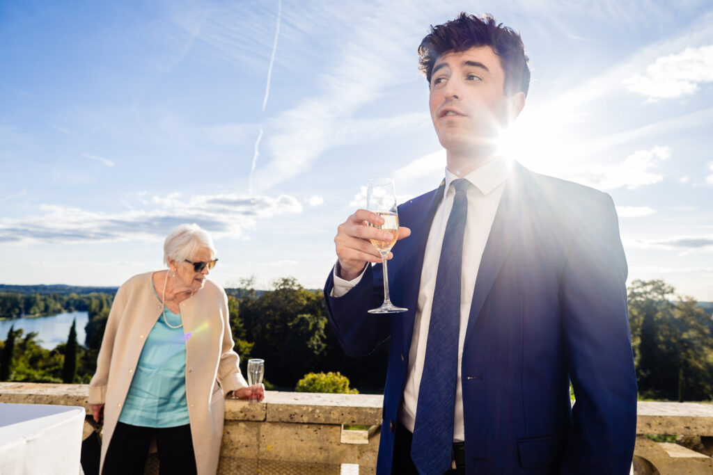 Un homme en costume tenant un verre se tient sur un balcon avec une vue panoramique tandis qu'une femme portant des lunettes de soleil regarde.