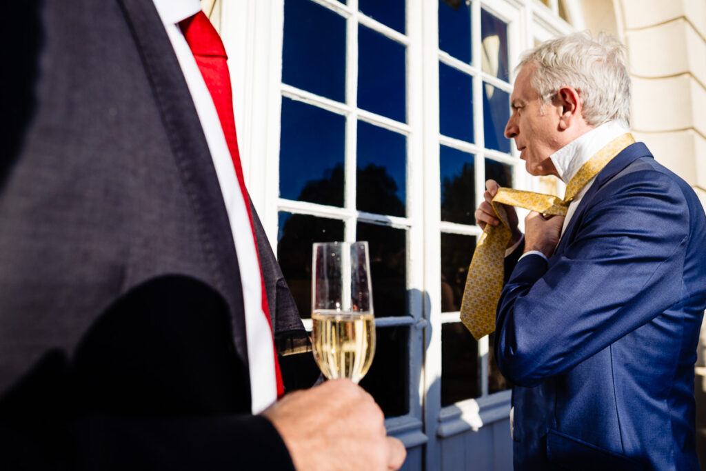 Deux hommes en costume se tiennent devant un bâtiment. L'un ajuste une cravate jaune tandis que l'autre tient une coupe de champagne. La lumière du soleil et les reflets des fenêtres créent une scène lumineuse et contrastée.