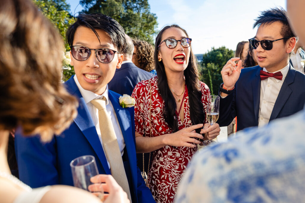 Des personnes en tenue de soirée et portant des lunettes de soleil sont rassemblées lors d'un événement en plein air, souriant et conversant tout en tenant des boissons.