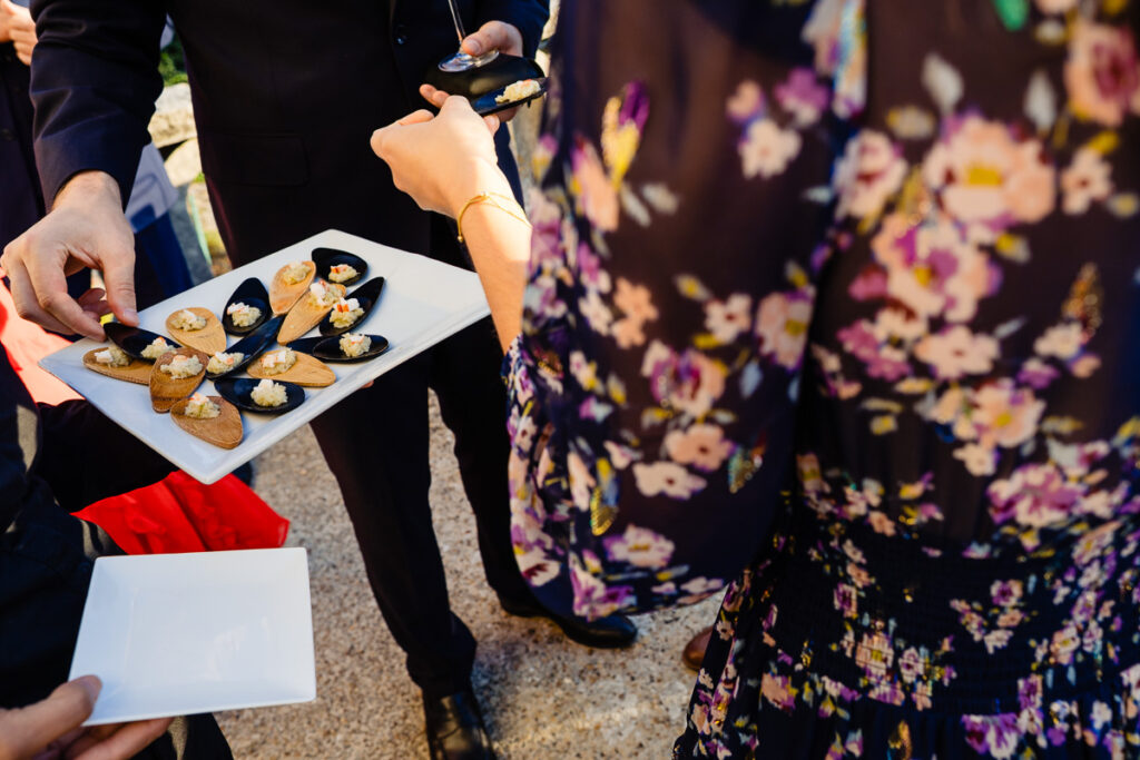 Une personne vêtue d'une robe fleurie et d'autres personnes se rassemblent autour d'un plateau contenant des amuse-gueules sur des cuillères noires, sur fond extérieur.