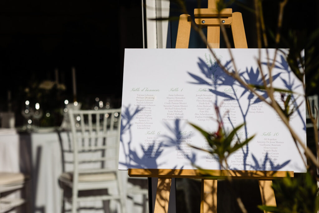 Un plan de table sur un chevalet est posé à l'extérieur, indiquant la répartition des tables. Les ombres des feuilles environnantes se projettent sur le tableau. Des chaises et des tables blanches avec leurs couverts sont visibles en arrière-plan.