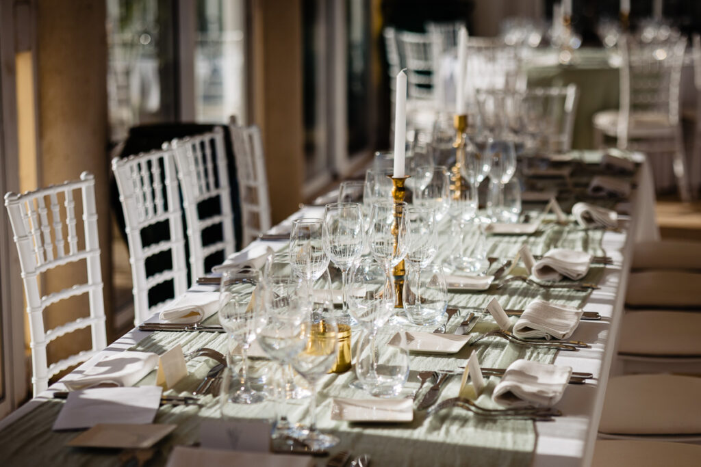 Une longue table à manger est élégamment dressée avec des chaises blanches, de la verrerie, des serviettes pliées, des couverts, des chandeliers et des petits marque-places, sous la lumière naturelle.