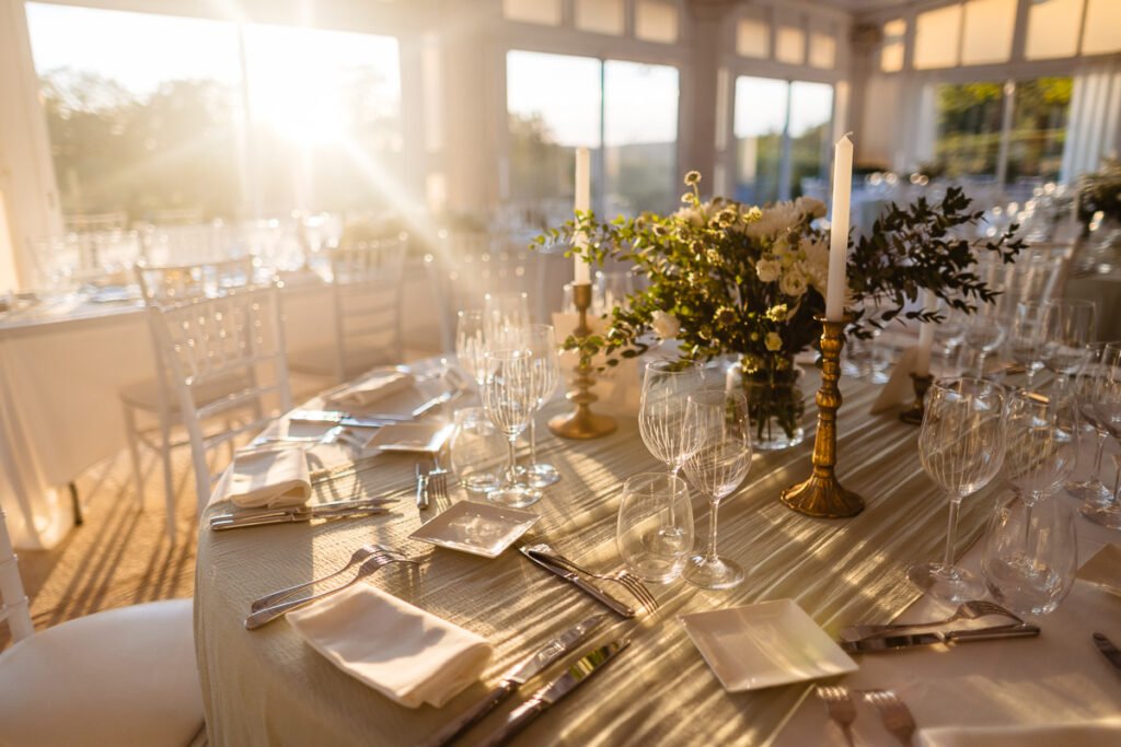 Une table ronde dressée pour un événement formel, avec verrerie, bougies et centre de table floral. La lumière du soleil filtre à travers de grandes fenêtres, illuminant la scène d'une douce lumière.