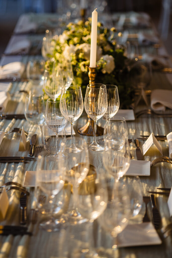 Une longue table à manger est élégamment dressée avec plusieurs verres à vin, assiettes, couverts et une bougie allumée dans un bougeoir. Un centre de table floral orne le centre.