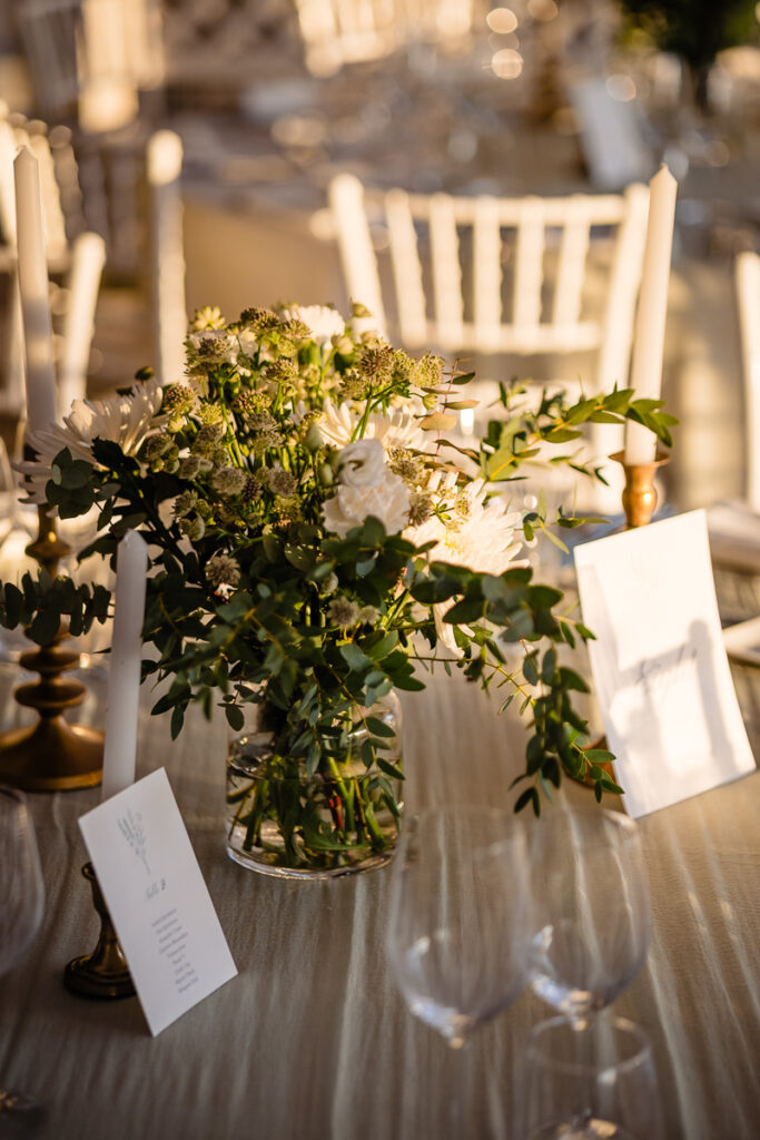 Un vase en verre avec des fleurs blanches et de la verdure sur une table dressée pour un événement. Des chaises blanches et une lumière douce et chaleureuse remplissent l'arrière-plan.