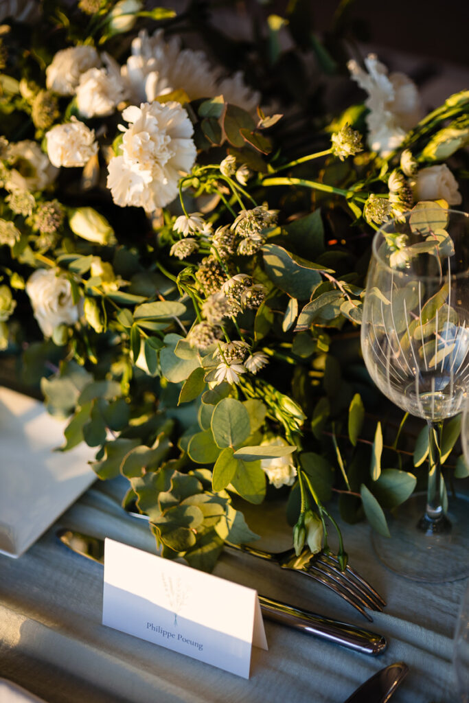 Décoration de table élégante avec des fleurs blanches, du feuillage vert, un verre à vin, des couverts et un marque-place sur une table ensoleillée.