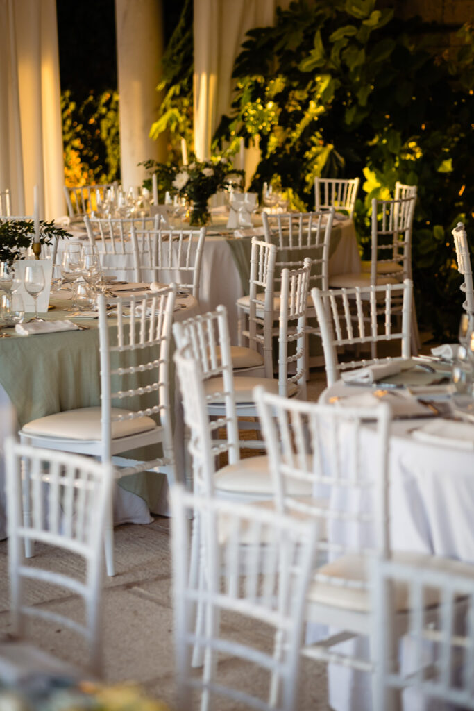 Des chaises blanches et des tables rondes nappées de blanc sont disposées pour un événement dans un espace ouvert. Une végétation luxuriante décore l'espace.