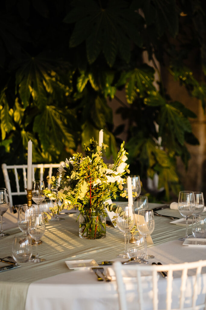 Une table ronde dressée pour un événement, comprenant des chaises blanches, une pièce maîtresse florale avec des bougies, de la verrerie et des couverts, avec de la verdure en arrière-plan.