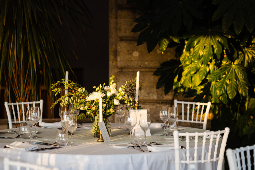 Une table ronde dressée pour un événement avec une nappe blanche, des chaises blanches, de la verrerie, des bougies blanches et un centre de table floral sous du feuillage.