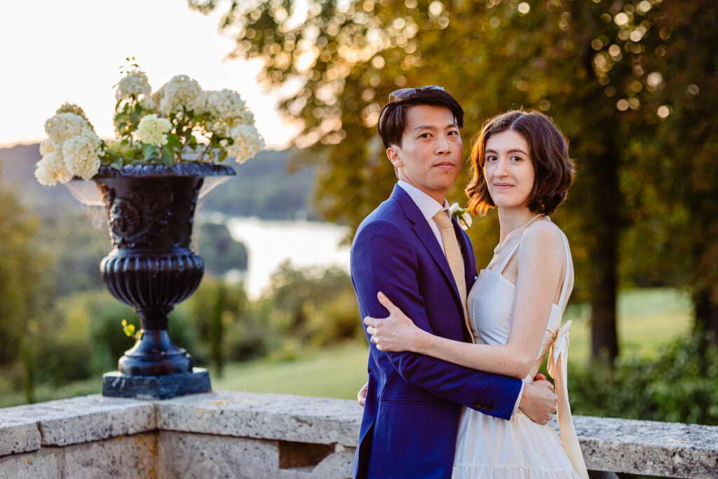 Un couple se tient ensemble à l'extérieur sur un balcon en pierre, entouré de verdure et d'un grand vase à fleurs, avec une vue panoramique en arrière-plan.