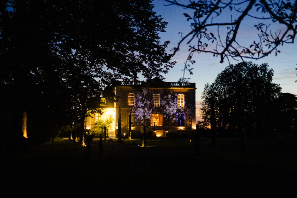Grand bâtiment avec fenêtres illuminées et silhouettes d'arbres au crépuscule.