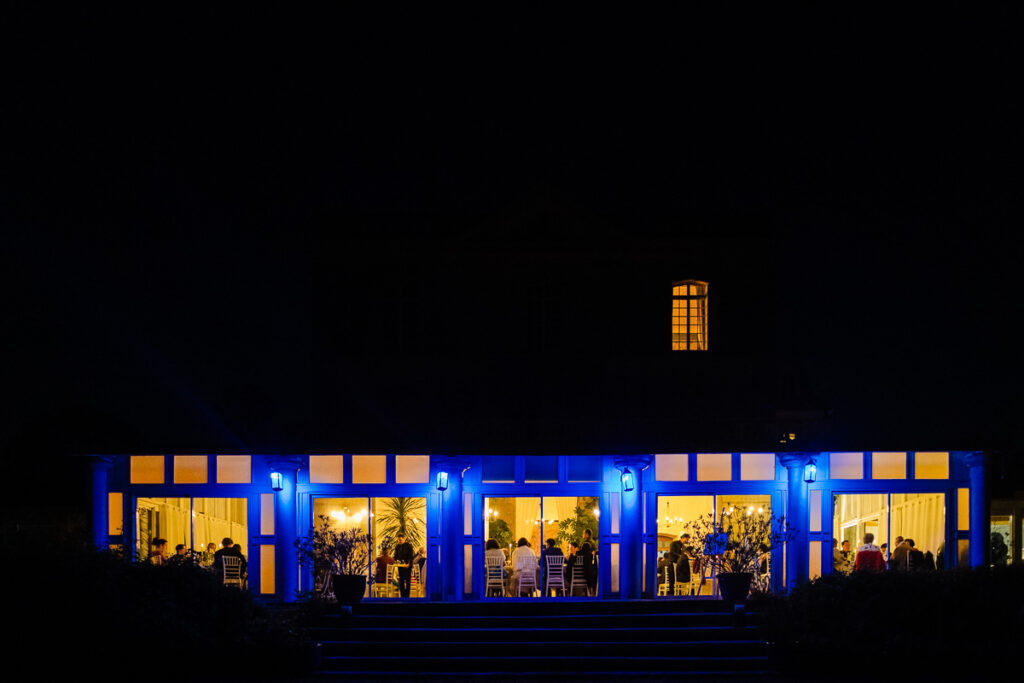 Un bâtiment lumineux, avec de grandes fenêtres et des personnes à l'intérieur, se détache sur un ciel nocturne sombre. Des lumières bleues illuminent l'extérieur.