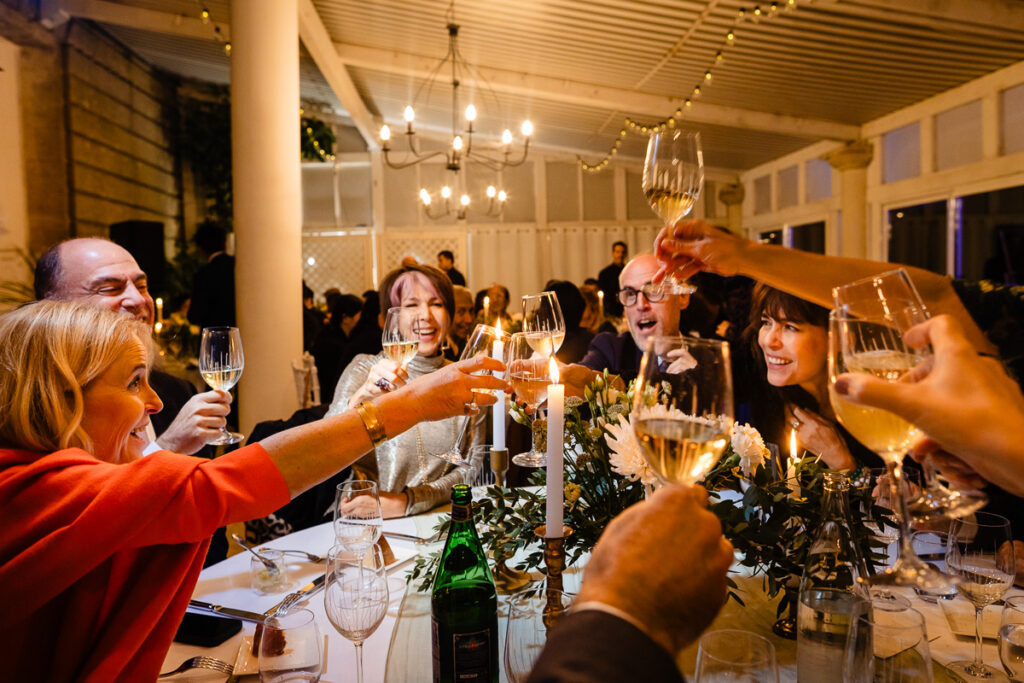 Groupe de personnes assises autour d'une table, levant leurs verres pour porter un toast, entourées de bougies et de compositions florales dans une pièce chaleureusement éclairée.