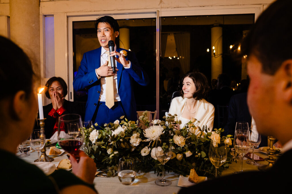 Un homme en costume bleu parle dans un microphone lors d'un événement officiel, entouré de personnes assises à une table avec des décorations florales et des verres.