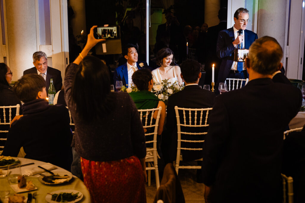 Un dîner où un homme prononce un discours devant des convives attablés. Un couple est assis, souriant, tandis que les invités prennent des photos.