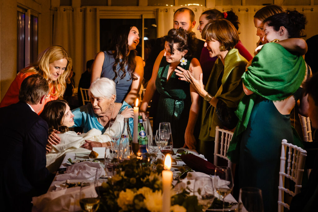 Un groupe de personnes se rassemble autour d'une table pour partager un moment convivial. Une femme âgée enlace une personne plus jeune, tandis que d'autres sourient et échangent. Bougies et fleurs décorent la table.