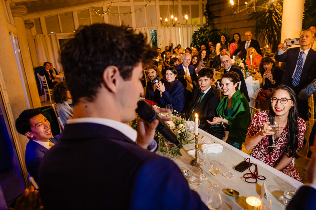 Une personne parle dans un microphone lors d'un événement officiel, s'adressant à des invités assis qui sourient et applaudissent, avec des bougies et des décorations visibles dans la salle.