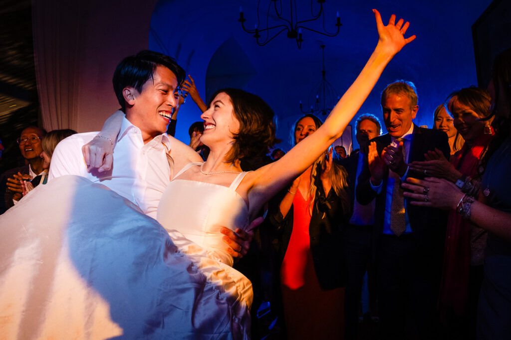 Un marié porte joyeusement sa mariée tout en célébrant avec une foule enthousiaste lors d'une réception de mariage.