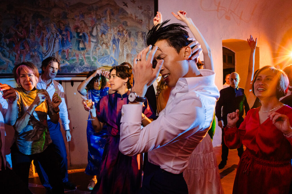 Des gens dansent énergiquement lors d'une fête intérieure animée, avec un éclairage coloré et une grande tapisserie murale en arrière-plan.