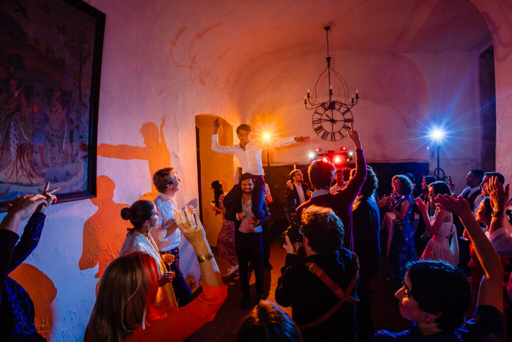 Les gens dansent et font la fête à l'intérieur, sous des lumières colorées. Un homme est porté sur les épaules d'un autre, entouré d'une foule en liesse. Une grande horloge est visible au mur.