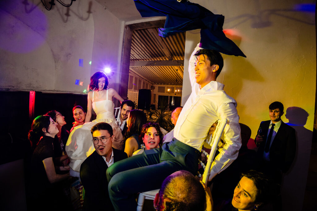 Des gens lors d'une fête animée, soulevant deux individus assis sur des chaises, avec un éclairage coloré créant une atmosphère festive.
