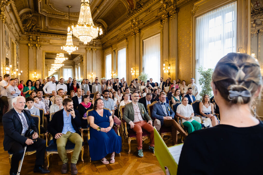 Un grand groupe de personnes est assis et debout dans une grande salle ornée de lustres, écoutant une femme parler au premier plan.