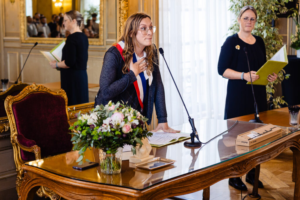 Une femme portant une écharpe tricolore parle sur un podium dans une salle formelle, avec des papiers sur le bureau et une autre femme debout à proximité tenant un dossier.