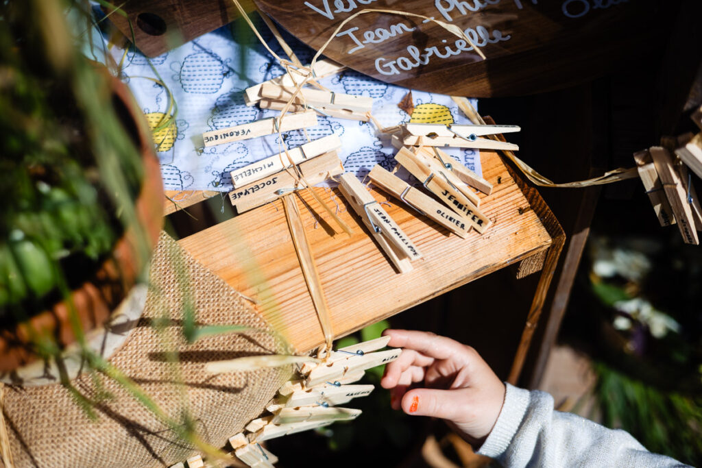 Une main se tend vers des pinces à linge attachées à une ficelle sur un présentoir en bois, chaque épingle étant étiquetée avec des noms ou des mots, entourée de plantes et de soleil.