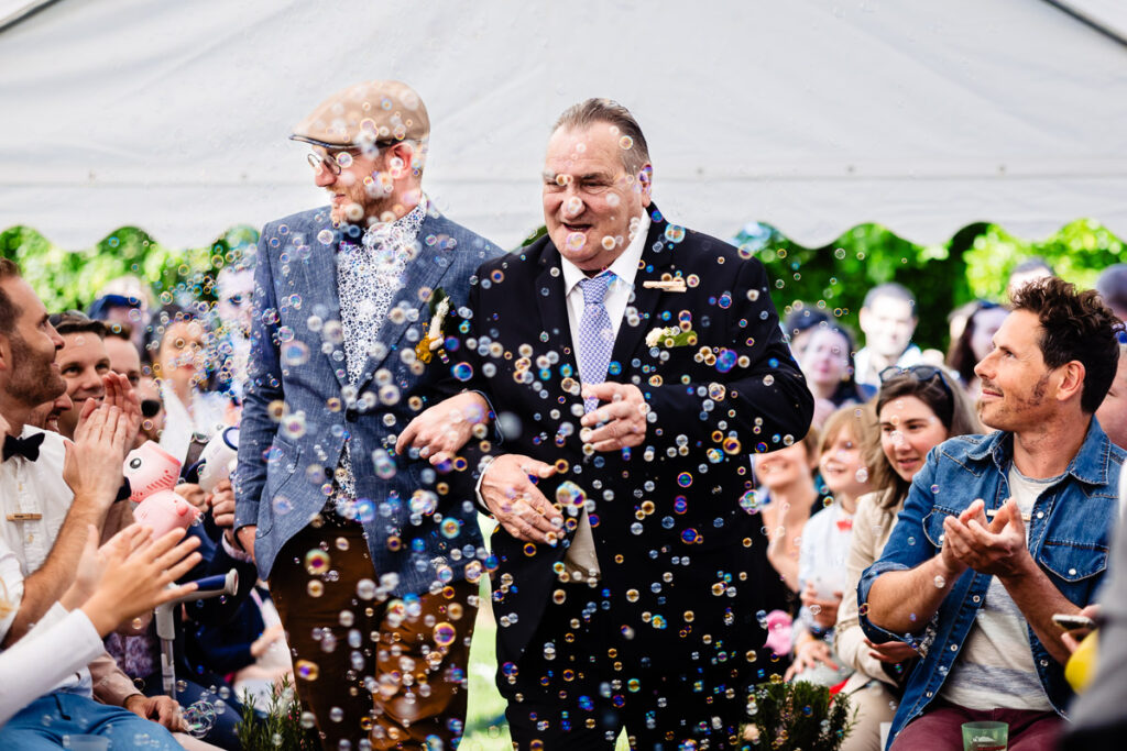 Un homme âgé souriant en costume marche bras dessus bras dessous avec un homme plus jeune dans une allée à l'extérieur, entouré de personnes et de bulles flottantes.