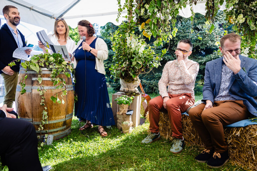 Lors d'un événement en plein air, un groupe de personnes se tient debout et s'assoit au milieu de la verdure et des balles de foin, certains s'essuyant les yeux tandis que d'autres lisent des journaux.