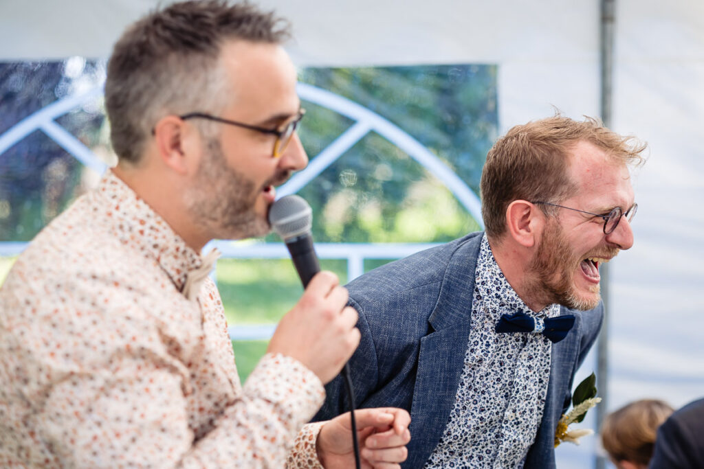 Deux hommes en chemises à motifs, l'un tenant un microphone et parlant, l'autre riant, se tiennent ensemble lors d'un événement en plein air sous une tente blanche.