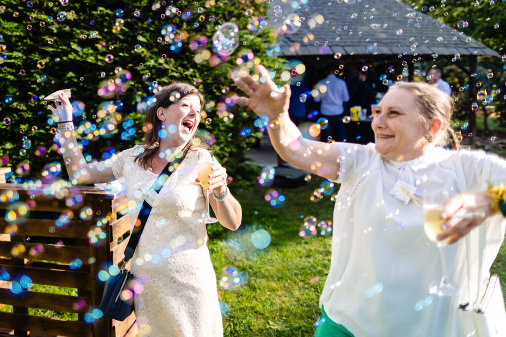 Deux femmes tenant des boissons sourient et tendent la main vers des bulles flottantes à l'extérieur lors d'un événement ensoleillé, avec de la verdure et des personnes en arrière-plan.
