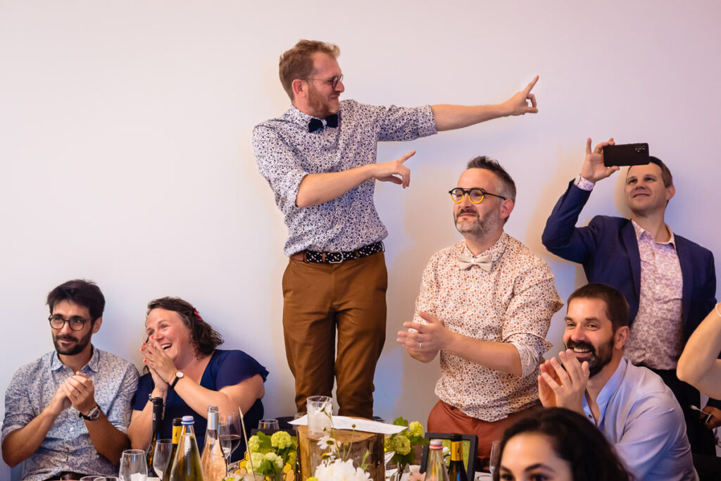 Un groupe de personnes à une table réagit avec des sourires et des applaudissements, tandis qu'un homme se tient debout sur une chaise et pointe du doigt tandis qu'un autre enregistre le moment avec un téléphone.