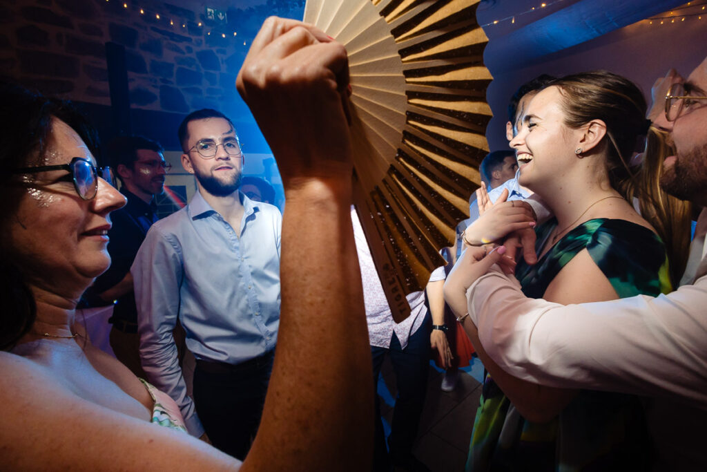Des gens dansent lors d'une fête sous des guirlandes lumineuses. Une femme au premier plan tient un grand éventail, tandis que d'autres sourient et interagissent en arrière-plan.