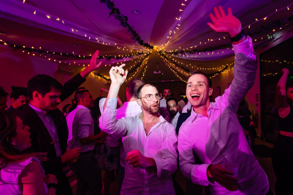 Un groupe de personnes danse et fait la fête sous des guirlandes lumineuses lors d'une fête, avec deux hommes au premier plan levant les bras et souriant.